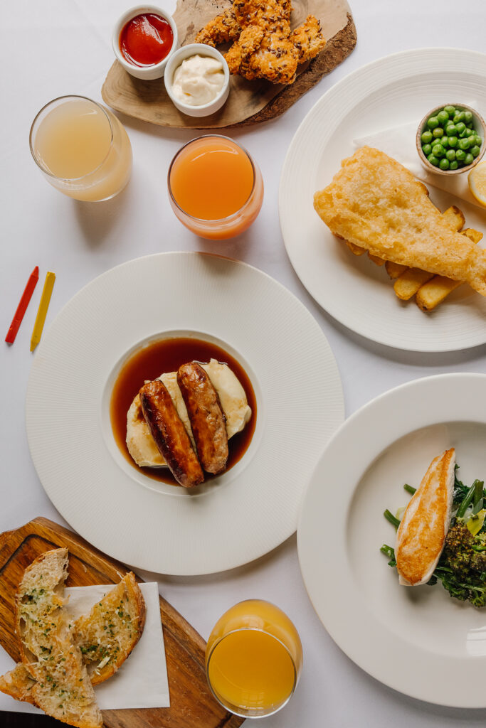 Small plates of fish and chips, sauage and mash, chicken and veg laid on a table with crayons